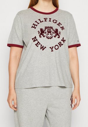 Osoba ubrana w szarą koszulkę z krótkim rękawem i spodnie z bordowym wykończeniem, z napisem „Hilfiger New York” oraz dwoma lwami wokół emblematu z tarczą.