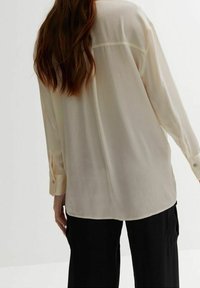 Woman with long brown hair wearing a loose, cream-colored blouse and black pants, standing against a plain light background.