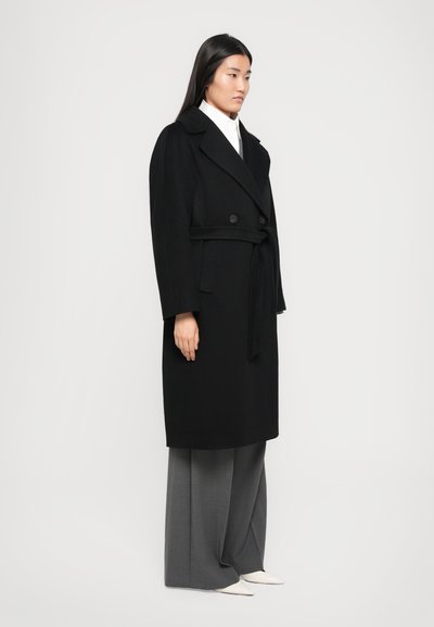 Black wool coat with a double-breasted front, wide collar, and a tie belt, paired with gray wide-leg trousers and white pointed shoes.