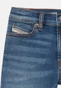 Blaue Jeans aus Denim mit einer Vordertasche und einem Logopatch, Kontrastnähten und einem Metallknopfverschluss. Der Stoff hat eine glatte, leicht strukturierte Oberfläche.