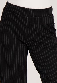 Zwarte broek met verticale strepen en een aangesloten tailleband, gemaakt van gladde stof, met subtiele metalen accenten in de strepen.