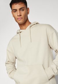 Beige hættetrøje lavet af blødt stof, med en frontlomme og snore i halsen. Glat tekstur og afslappet pasform.
