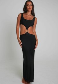 Frau in einem ärmellosen schwarzen Maxikleid mit Ausschnitten und einem silbernen Ringdetail in der Taille, barfuß vor einem schlichten Hintergrund stehend.