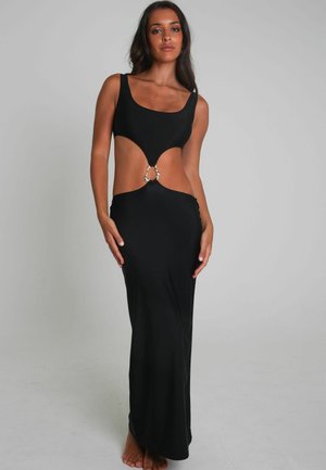 Vrouw in een mouwloze zwarte maxi-jurk met uitsnijdingen en een zilveren ringdetail op de taille, staand op blote voeten tegen een effen achtergrond.