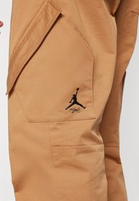 Poche de pantalon beige avec logo Jumpman noir brodé et texte "Flight", comprenant un rabat et des détails de coutures visibles.
