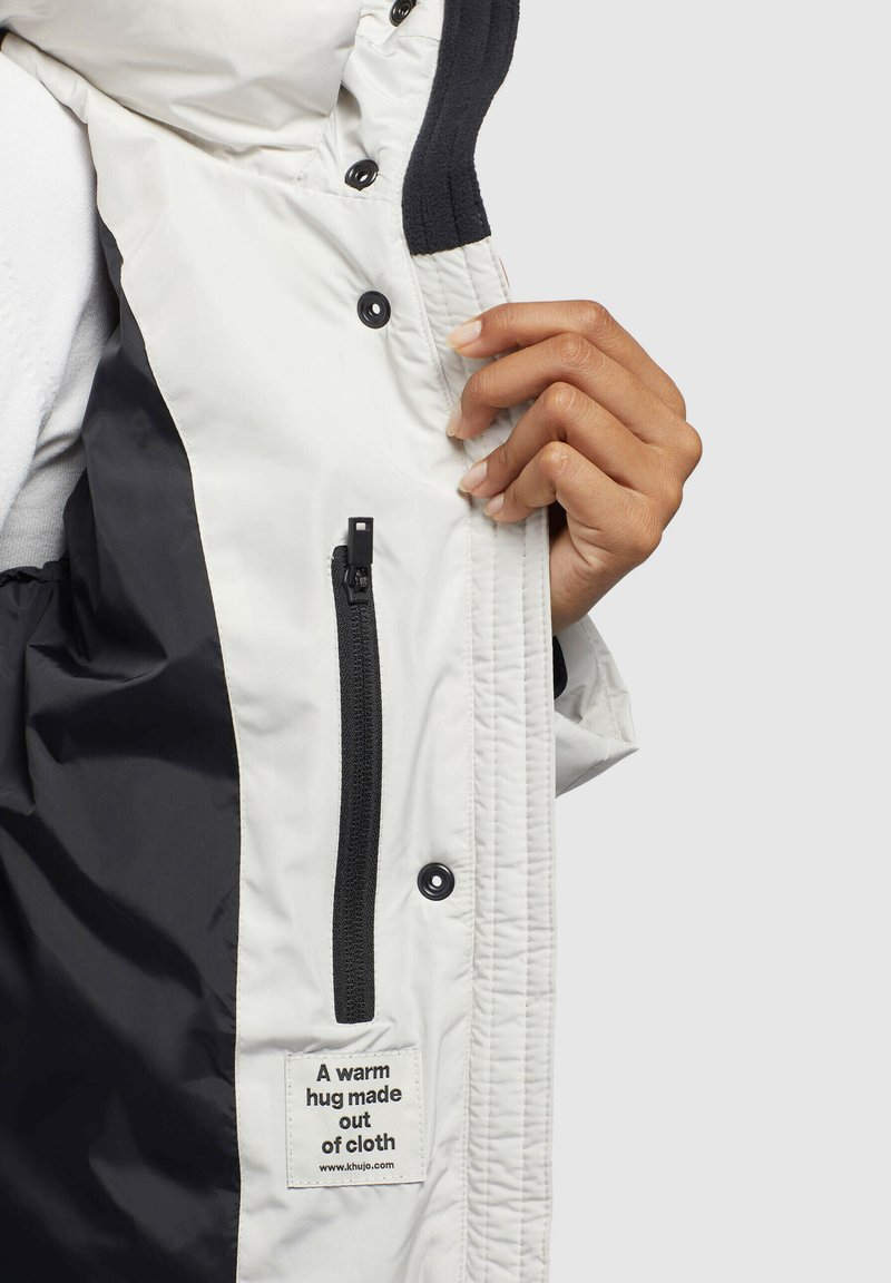 Gesteppten Jacke in Weiß mit einem schwarzen Innenfutter, ausgestattet mit einer schwarzen Reißverschlusstasche, Druckknöpfen und einem Textillabel, das sagt: "Eine warme Umarmung aus Stoff."