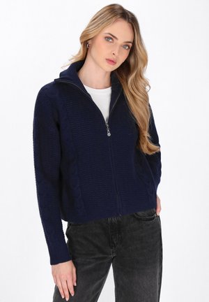 Maglione zip-up blu navy con un design a trecce testurizzato, dotato di un colletto alto e maniche lunghe. Abbinato a jeans grigio scuro.