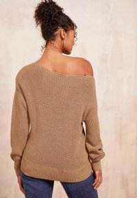 Übergroßer, schulterfreier Pullover aus beigem Strick, mit gerippten Bündchen und Saum. Kombiniert mit dunkelblauen Jeans. Strukturierter Stoff sichtbar.