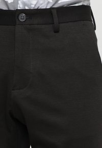 Pantalon noir en tissu tissé, avec une texture lisse, une ceinture standard avec passants pour ceinture, et une fermeture par bouton à l'avant.