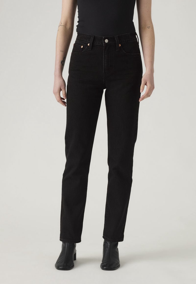 Personne portant un jean noir taille haute à jambes droites, un haut noir sans manches et des bottines noires, debout devant un fond clair uni.