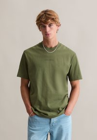Oliwkowa bawełniana koszulka z krótkimi rękawami, okrągłym dekoltem i subtelnym logo na przodzie, zestawiona z jasnoniebieskimi dżinsami.