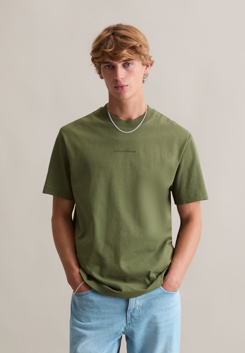 Oliwkowa bawełniana koszulka z krótkimi rękawami, okrągłym dekoltem i subtelnym logo na przodzie, zestawiona z jasnoniebieskimi dżinsami.