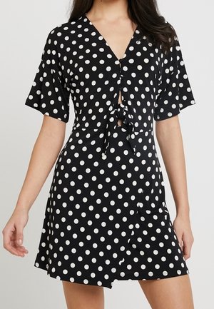 Robe noire à pois blancs, avec un décolleté en V, un détail de nœud à l'avant, des manches courtes et un ourlet évasé. Des boutons ornent le devant.