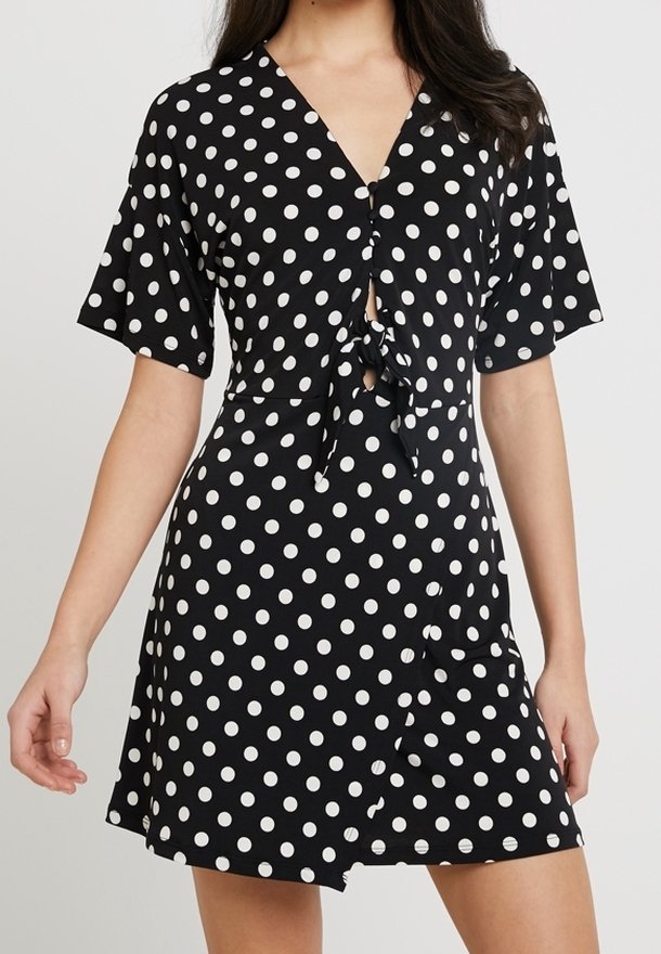 Robe noire à pois blancs, avec un décolleté en V, un détail de nœud à l'avant, des manches courtes et un ourlet évasé. Des boutons ornent le devant.