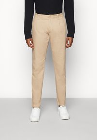 Beige chinos with a straight cut, featuring front pleats. Button closure at the waist, paired with white sneakers. Smooth fabric texture.