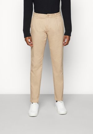 Beige chinos with a straight cut, featuring front pleats. Button closure at the waist, paired with white sneakers. Smooth fabric texture.