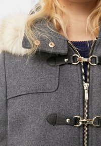 Manteau en laine grise avec des finitions en fausse fourrure, doté d'une fermeture éclair, de fermoirs décoratifs et d'une finition texturée. Les accents incluent du matériel doré.