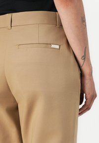 Pantaloni beige con una texture liscia, presentano una tasca posteriore e un sottile dettaglio con etichetta. La cintura è piatta e le cuciture sono minime e ordinate.