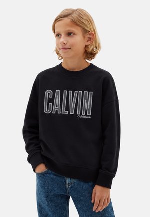 Bambino biondo che indossa una felpa nera Calvin Klein con grande logo e jeans blu, in piedi con una mano in tasca.