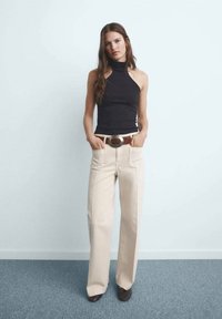 Black sleeveless top with high neckline, beige wide-leg trousers, and brown belt with a large circular buckle; standing against a light background.