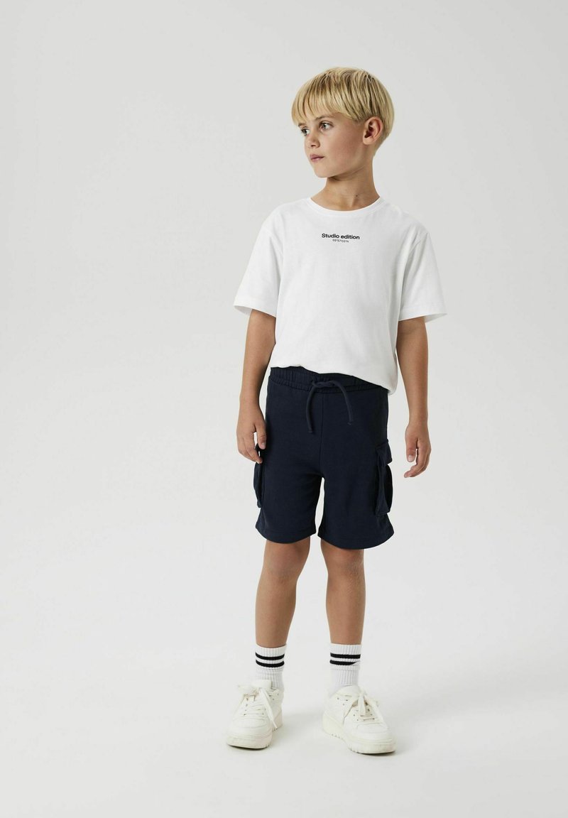 T-shirt blanc avec texte noir, short bleu marine avec poches latérales, baskets blanches et chaussettes rayées noires et blanches. Conception simple et décontractée.