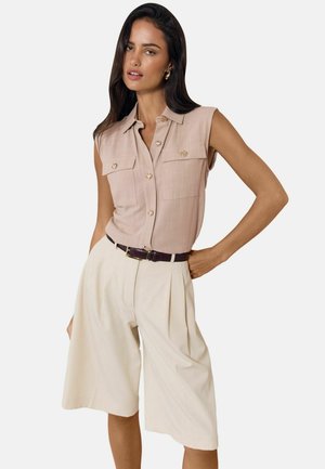 Žena nosi bezové tielkové košeľu na gombíky s prednými vreckami a krémové široké krátke nohavice po kolená s čiernym opaskom.