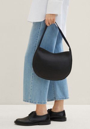 Schwarze Leder-Schultertasche mit geschwungener Form, Griff und glatter Textur, kombiniert mit hellblauen weit geschnittenen Jeans und schwarzen Schuhen.