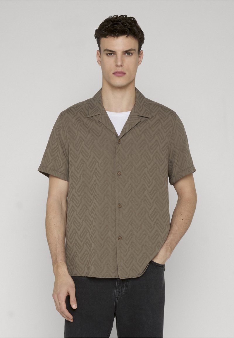 Jeune homme aux cheveux courts et bouclés, portant une chemise marron à motifs à manches courtes boutonnée sur un débardeur blanc, debout avec une main dans la poche.