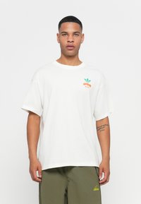 Hvid bomulds t-shirt med rund halsudskæring og korte ærmer. Har et grønt og orange "Adidas" logo grafik på venstre bryst.