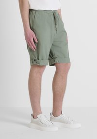 Pantaloni corti verdi con una vita elastica e un orlo arrotolato con dettaglio a bottone. Realizzati in tessuto leggero. Abbinati a sneakers bianche.