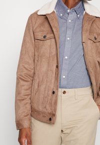 Chaqueta de ante marrón con cuello de borrego sintético crema, que presenta dos bolsillos en el pecho y cierre de botones. Llevada sobre una camisa azul abotonada.