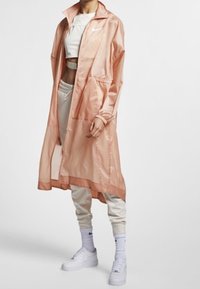 Persikofärgad, lätt nylon långjacka med hög krage, framfickor och transparent design, bärs över en vit crop top och sweatpants.