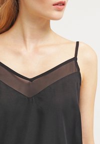 Top negro sin mangas con escote en V que presenta detalles de malla transparente y tirantes finos ajustables. Textura suave y tejido ligero.