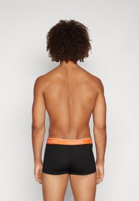 Black boxer briefs with an orange Lacoste logo on the waistband, made from a smooth, stretchy fabric. View from the back.