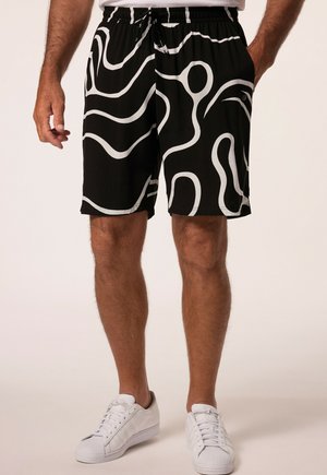 Sorte shorts med hvide abstrakte bølgede linjer båret af en person, der står, kombineret med hvide sneakers og en hvid skjorte.