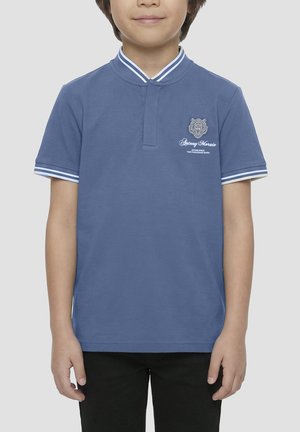 Dijete koje nosi plavu polo majicu kratkih rukava s bijelim prugastim ovratnikom i manžetama te izvezenim logotipom na lijevoj strani prsa.