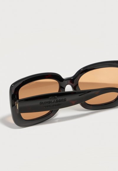Μαύρα ορθογώνια γυαλιά ηλίου με καφέ φακούς που ακουμπούν σε λευκή επιφάνεια, με την ένδειξη «THE SUNGLASSES» και το όνομα της μάρκας στο μπράτσο.