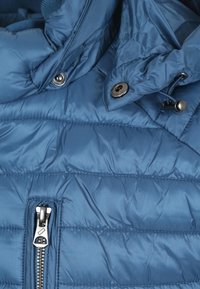 Blauwe geïsoleerde jas met een doorgestikt ontwerp, voorzien van een zilveren rits en metalen drukknoopjes. Gladde, glanzende stof met een zachte textuur.