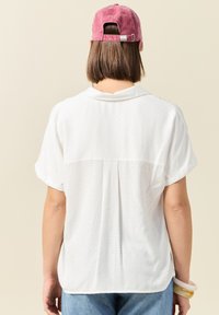 Camisa blanca de manga corta con un patrón texturizado, detalle de pliegue en la parte posterior y un corte relajado, combinada con jeans de color azul claro y una gorra rosa.