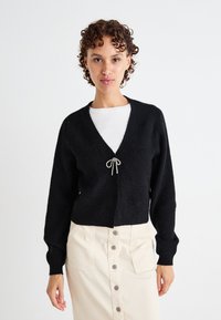 Cardigan negru tricotat, cu decolteu în V, mâneci lungi și închidere frontală cu un laț gri deschis. Purtați peste o cămașă albă și o fustă bej.