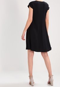 Černé šaty s krátkými rukávy v kapkovém střihu, A-line tvar, hladká textura, délka ke kolenům, v kombinaci s béžovými podpatky. Jednoduchý a minimalistický design.