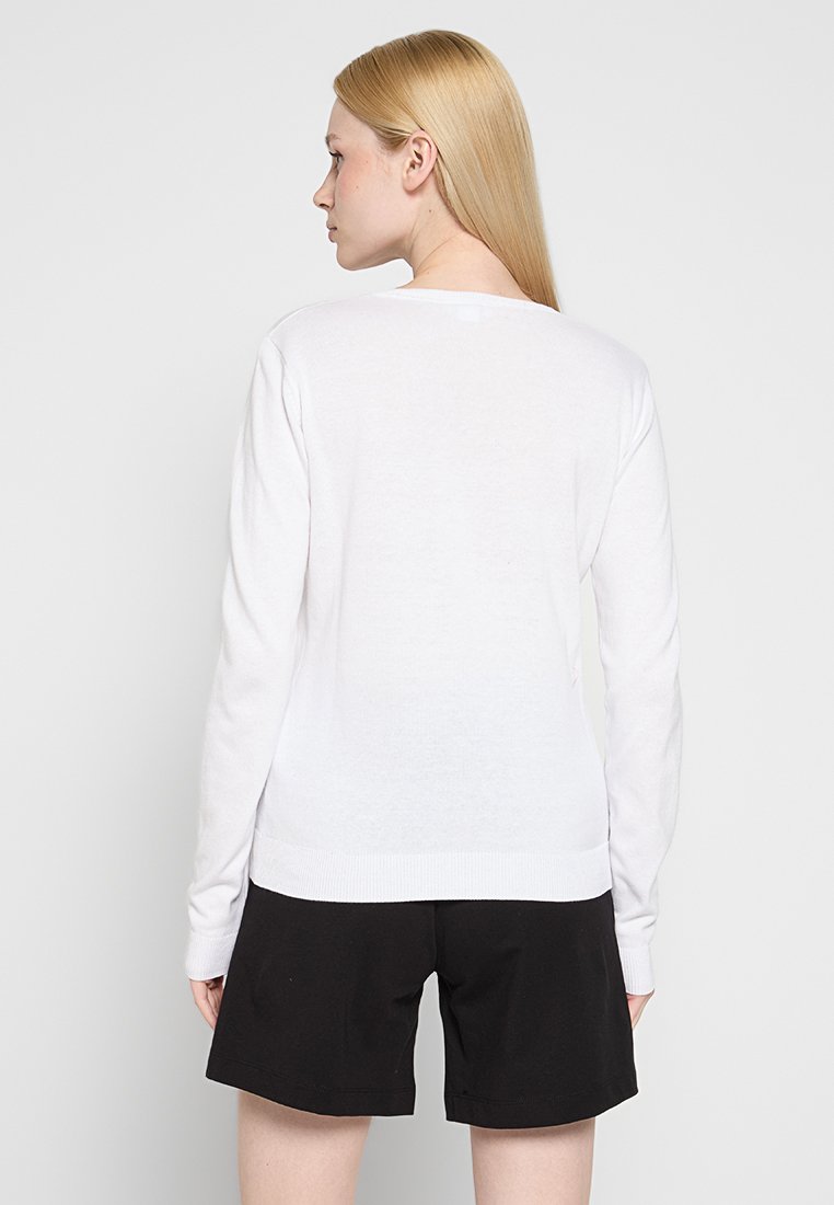 Frau mit glattem, blonden Haar, die ein weißes Langarmshirt und schwarze Shorts trägt und mit dem Rücken zu einem schlichten, hellen Hintergrund steht.