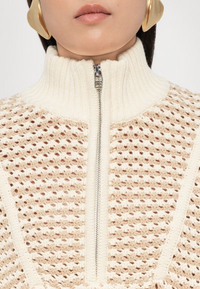 Jersey de punto que presenta un alto cuello canalé, cierre de cremallera y un patrón texturizado en beige con una banda de crema en contraste a lo largo de los bordes.