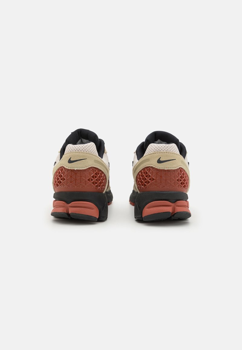 Buty biegowe Nike z beżową cholewką, czarnymi akcentami, teksturowaną czerwoną piętą oraz wyściełaną czarną podeszwą. Charakterystyczne geometriczne wzory na pięcie.