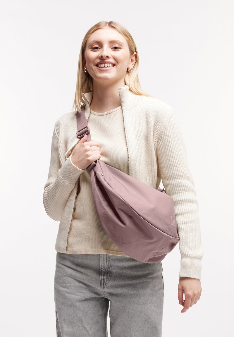 Borsa a tracolla in morbido tessuto rosa chiaro, con forma curva e tracolla regolabile. Il modello indossa un maglione beige a coste e jeans grigi chiari.