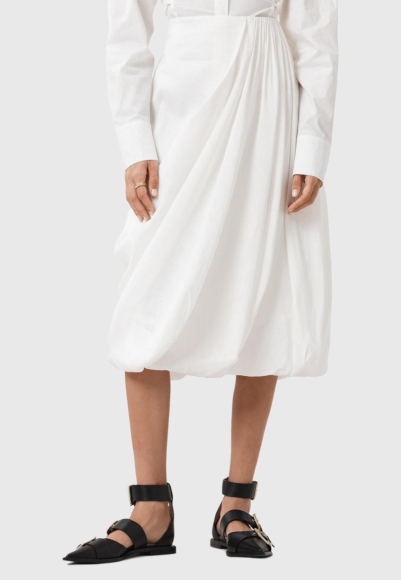 Biała, marszczona spódnica z gładkiego bawełnianego materiału, zestawiona z czarnymi płaskimi sandałami z zapięciem na klamrę.