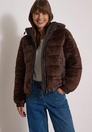 Femme portant une veste matelassée en peluche marron par-dessus une chemise blanche et un jean bleu, souriante, une main dans la poche, sur un fond uni.