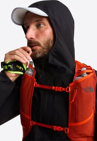 Jasnopomarańczowa kamizelka nawadniająca z siateczkowymi kieszeniami i paskami, połączona z czarną kurtką z kapturem. Kamizelka wyposażona jest w dwie butelki na wodę i elementy odblaskowe.