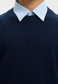 Pull en maille bleu marine avec un col rond porté sur une chemise à col rayée bleu clair, comportant un détail de bouton au niveau du col.