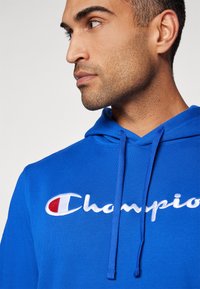 Kék kapucnis pulóver zsinórokkal, fehér hímzett "Champion" logóval. Az anyag lágy és sima textúrájú. Részletes közeli kép.